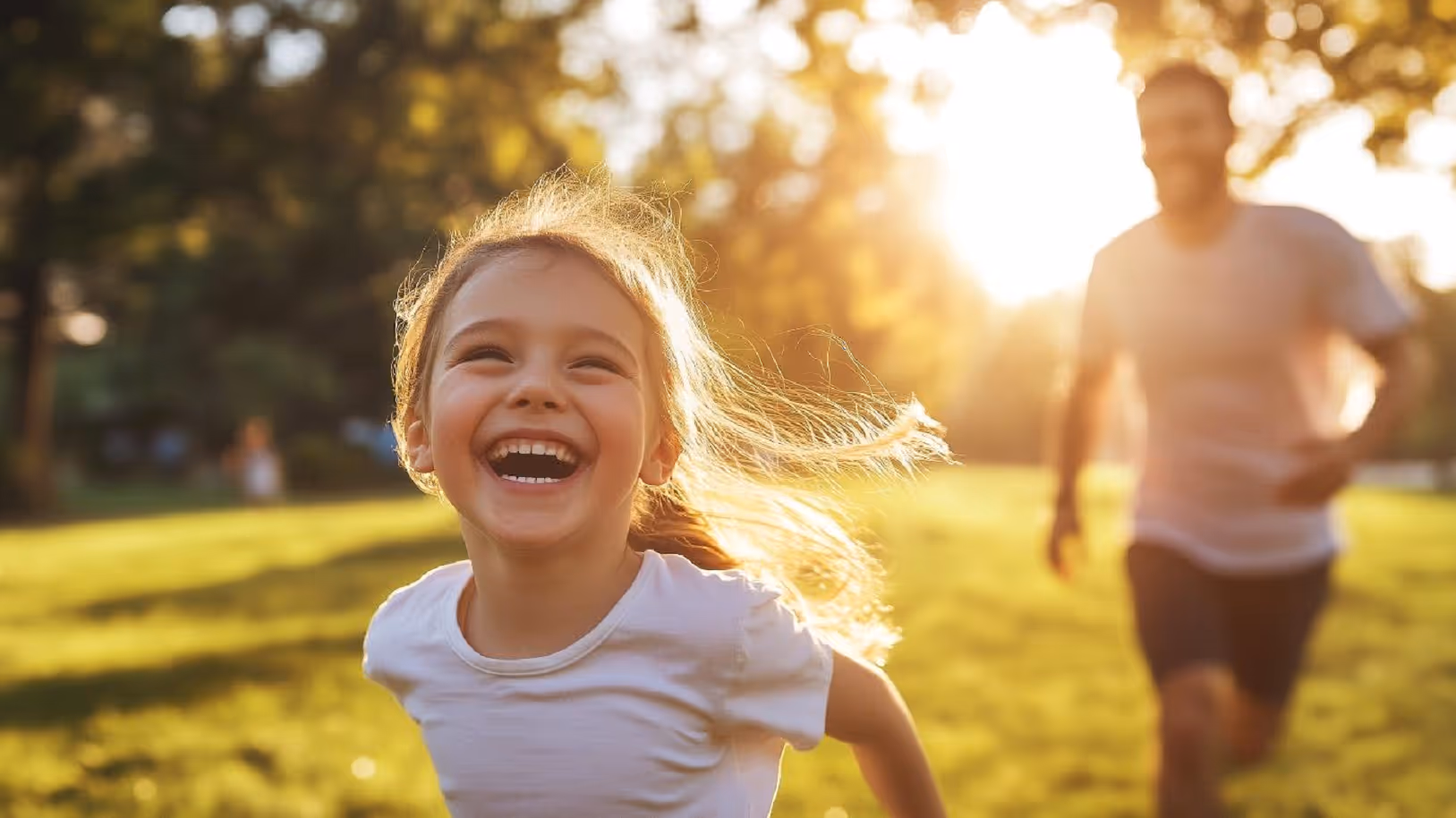 A joyful young girl running through a sunlit park, laughing with her hair blowing in the breeze. Her father follows behind her in the background.