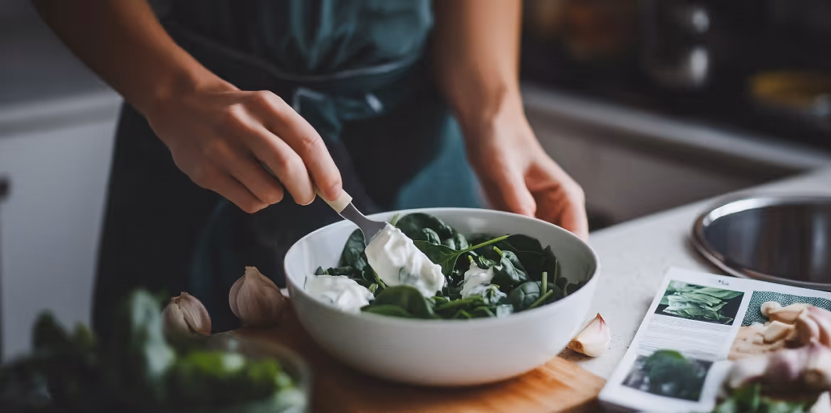 Close-up of a person preparing a bowl of spinach topped with yogurt, with garlic cloves