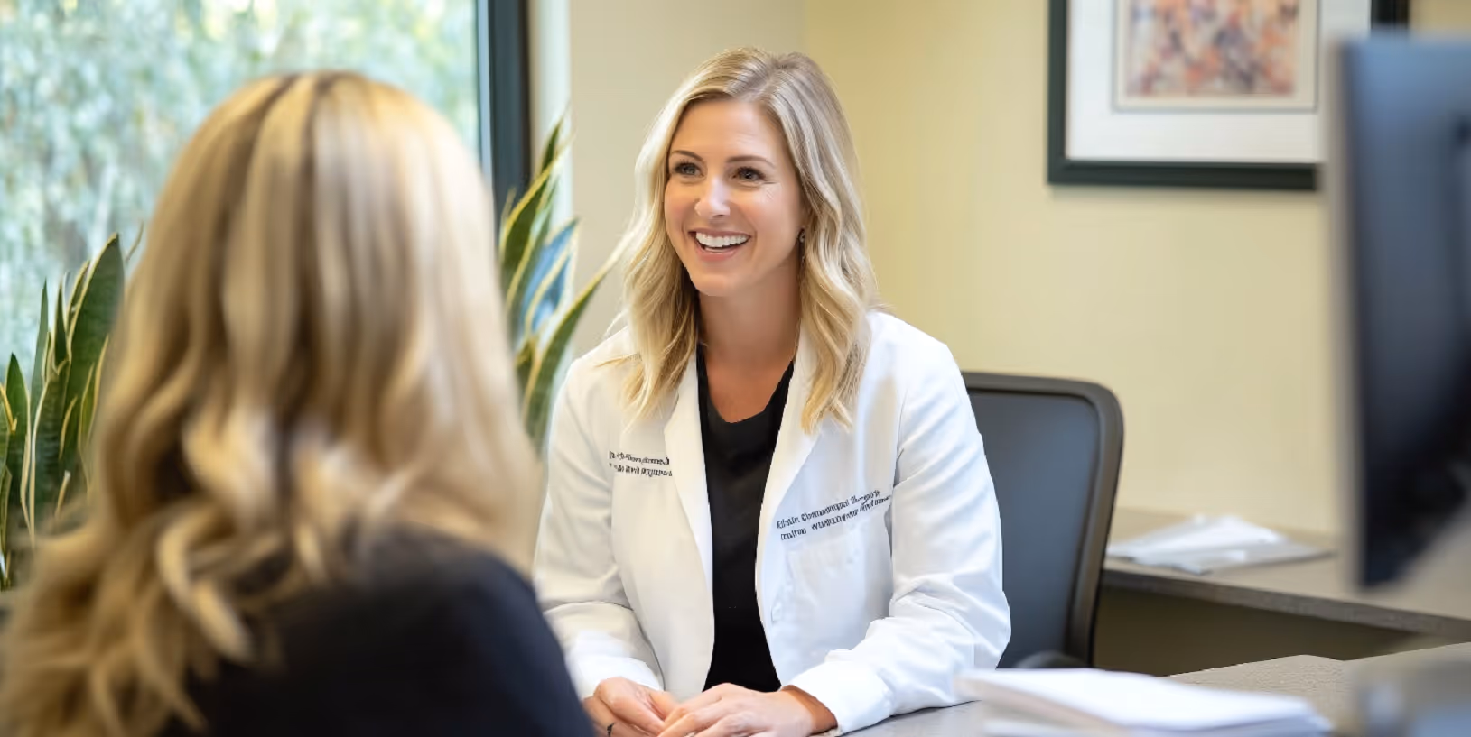A female doctor in a white coat smiling while talking to a patient across the desk in a bright medical office