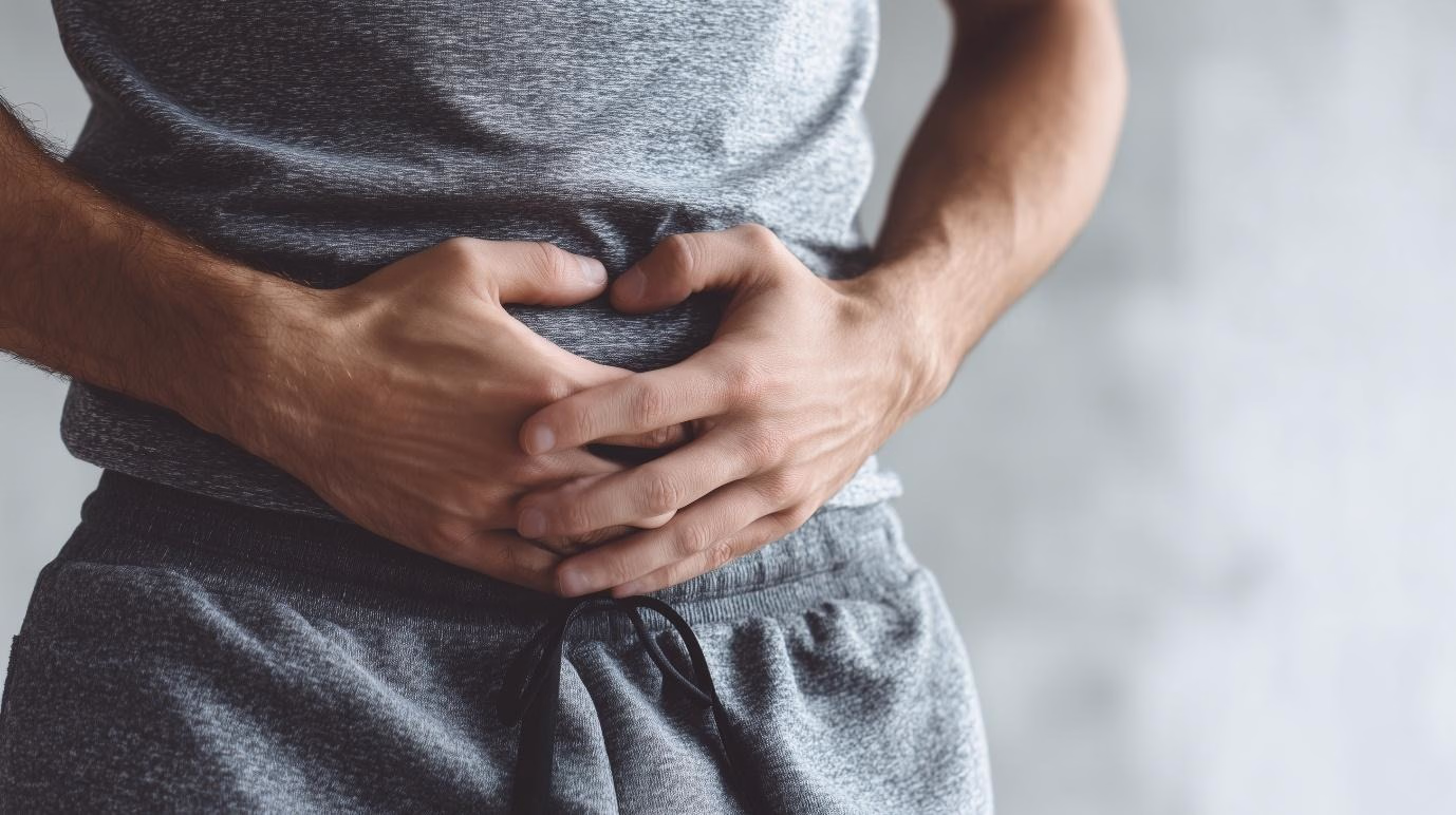 Close-up of a man’s midsection wearing grey athletic clothes, with both hands placed over his stomach, suggesting discomfort or abdominal pain.
