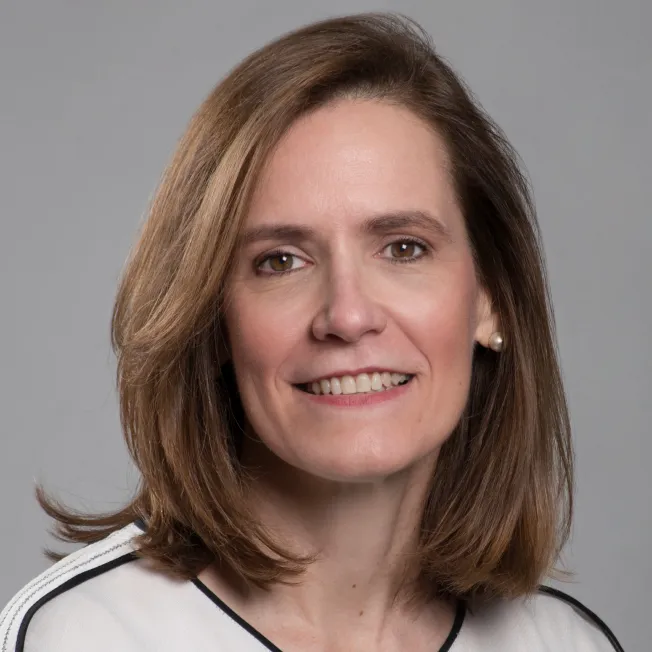 Portrait of a smiling woman with shoulder-length brown hair, wearing a white top with black trim and pearl earrings, against a gray background.