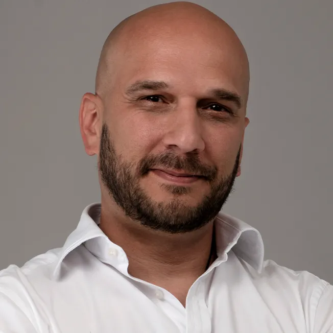 Portrait of a man with a neatly trimmed beard wearing a white shirt against a gray background.