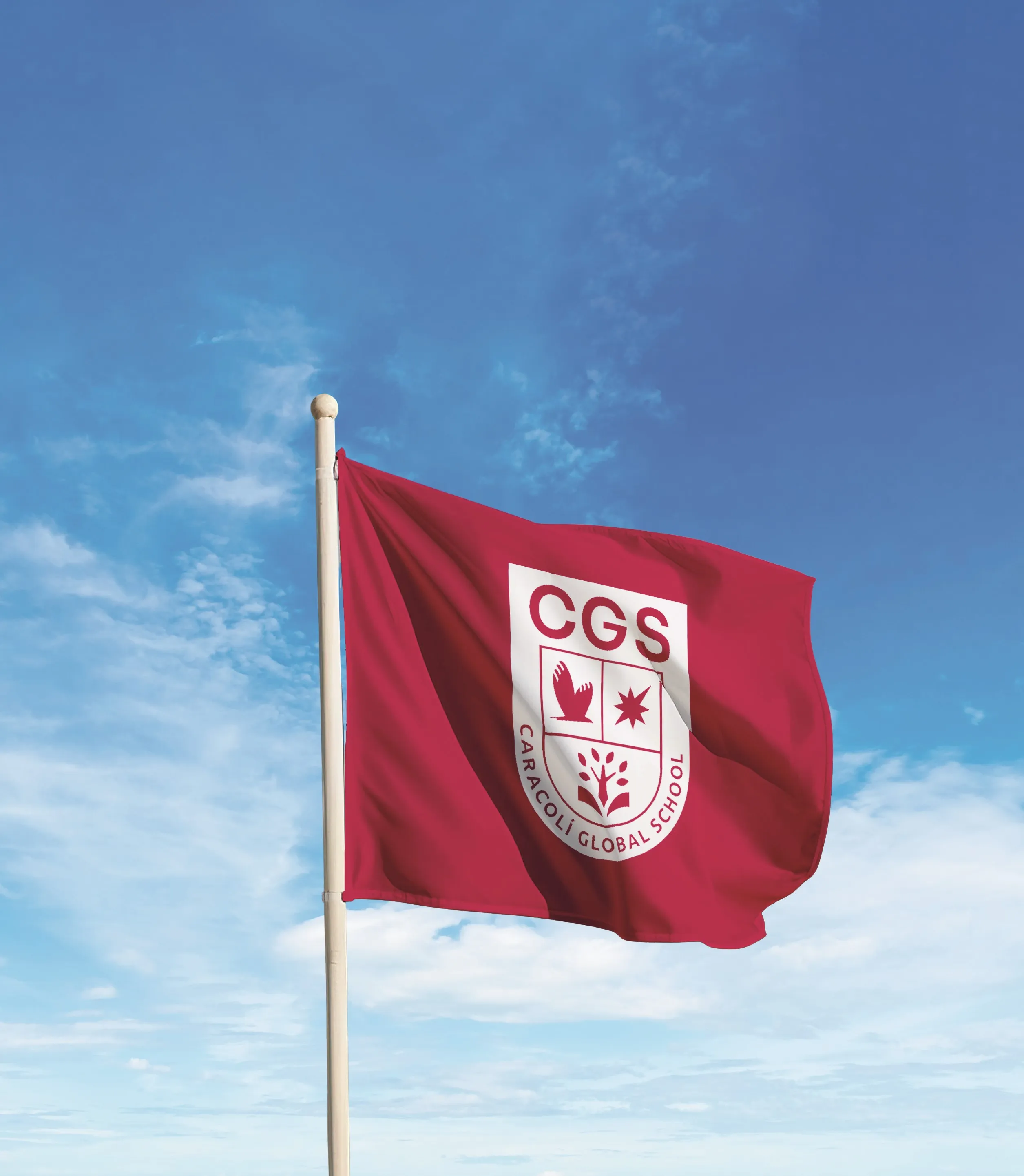 Red flag with a white shield emblem featuring a bird, a star, a tree, and the text 'CGS Caracoli Global School' waving against a blue sky with scattered clouds.