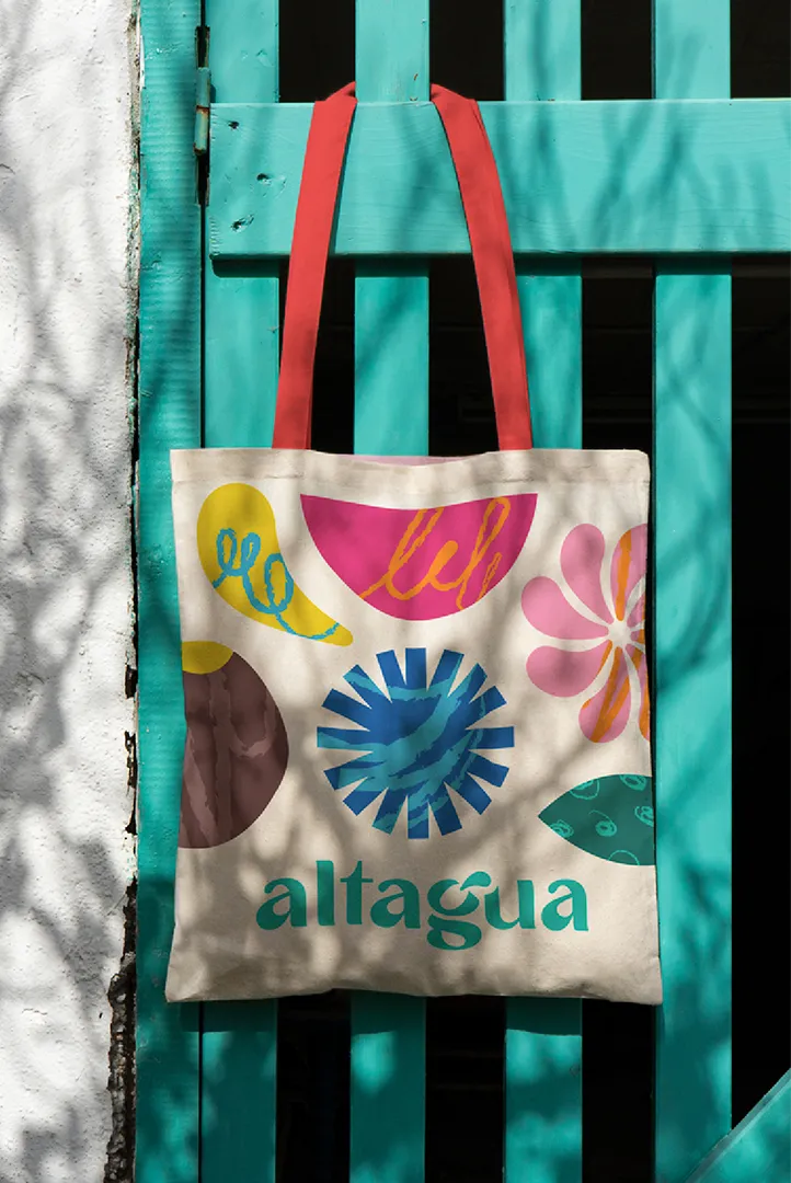 Canvas tote bag with colorful abstract shapes and the word 'altagua' hanging on a turquoise wooden fence.