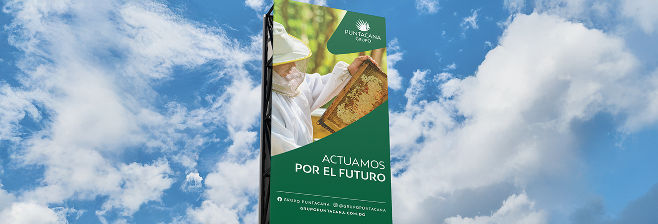 Tall vertical green billboard against blue sky with clouds showing a beekeeper holding a honeycomb and text 'Actuamos por el futuro' from Grupo Puntacana.