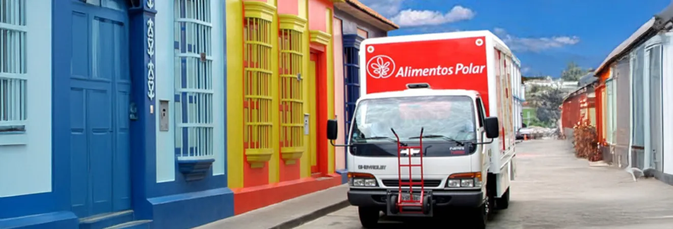 White delivery truck with red 'Alimentos Polar' logo parked on a street lined with colorful buildings under a blue sky.
