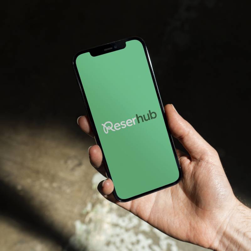 Hand holding a smartphone displaying a green screen with the Reserhub logo.