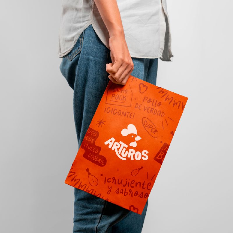 Person holding an orange paper bag with white logo and Spanish text for 'Arturos', describing chicken products.