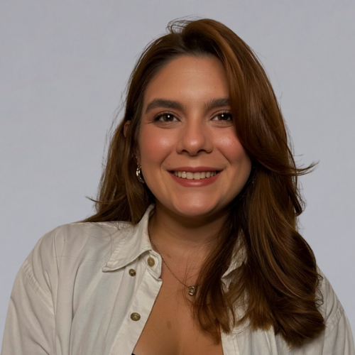 Smiling young woman with brown hair wearing a white shirt and gold hoop earrings against a plain light gray background.