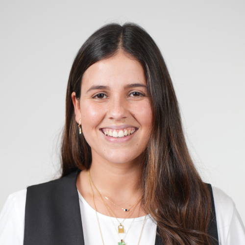Smiling woman with long brown hair wearing a white top, black vest, and gold necklaces.