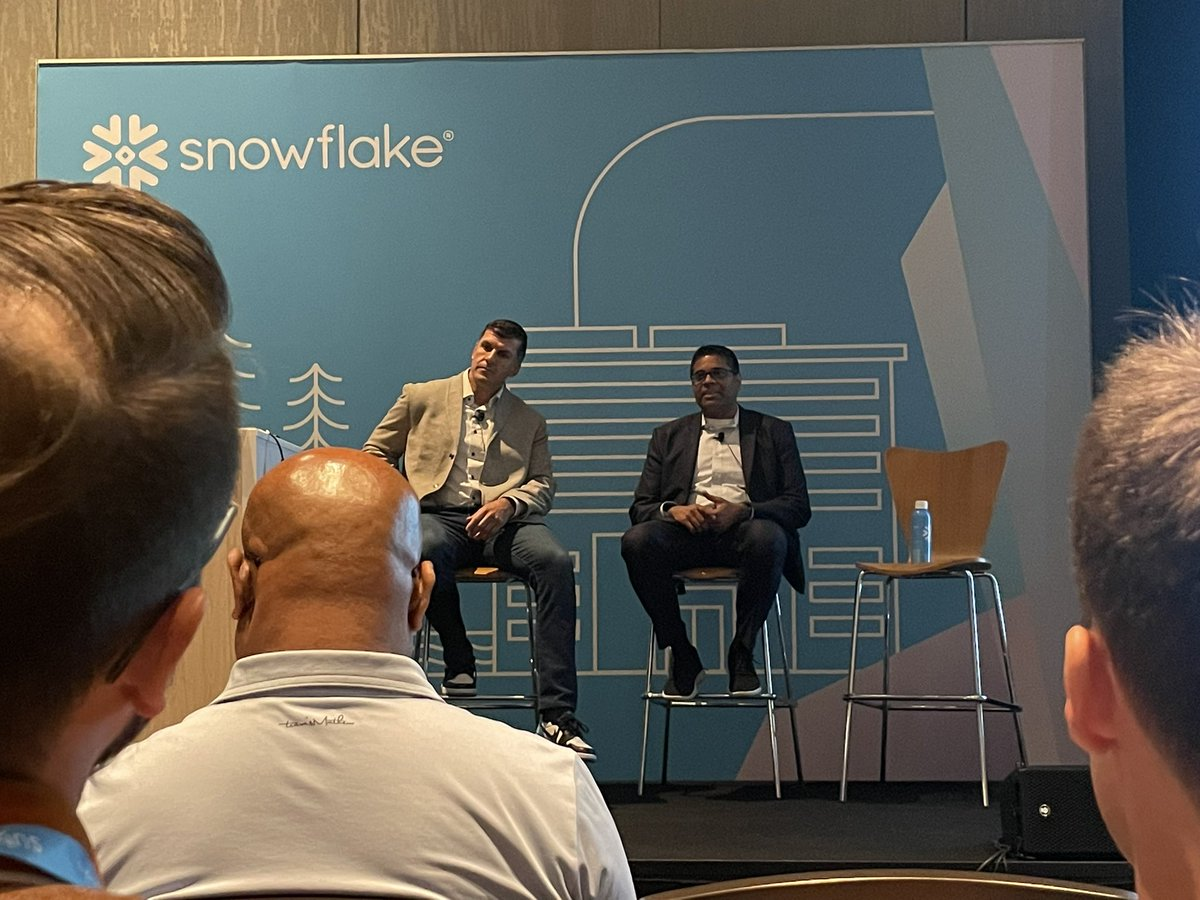 Two men sitting on stools in front of a blue sign that says Snowflake.