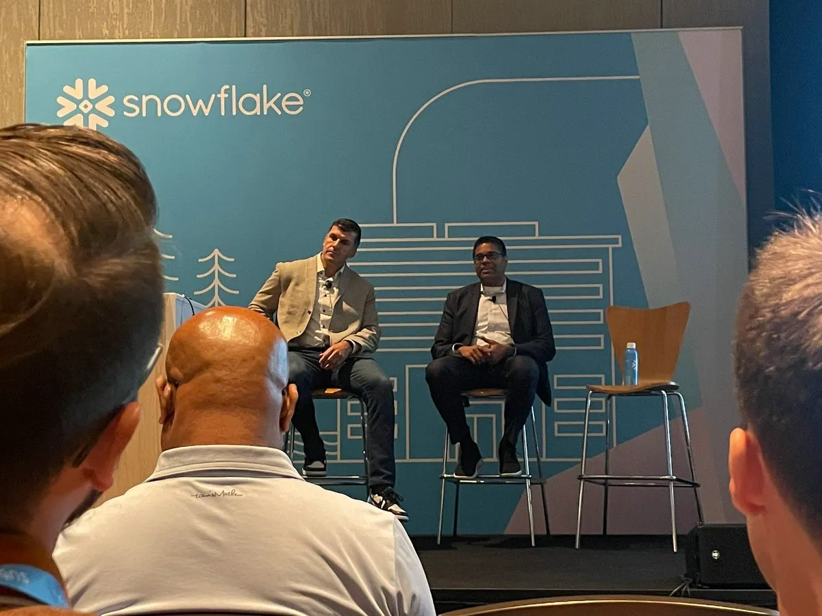 Two men sitting on stools in front of a blue sign that says Snowflake.