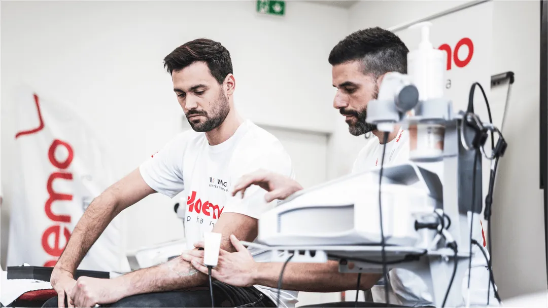 Ein Mann erhält eine physiotherapeutische Behandlung am Arm mit einem modernen medizinischen Gerät, betreut von einem Therapeuten, beide tragen Haemo Pharma T-Shirts.