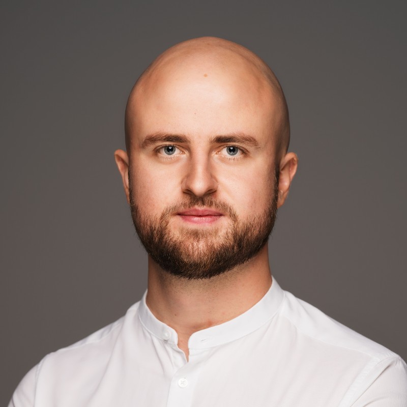 Mężczyzna z ogoloną głową i brodą w białej koszuli na jednolitym szarym tle.