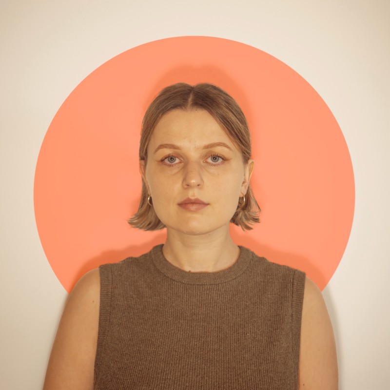 Portret kobiety z krótkimi blond włosami, ubraną w brązowy sweter bez rękawów, na tle pomarańczowego koła na jasnej ścianie.