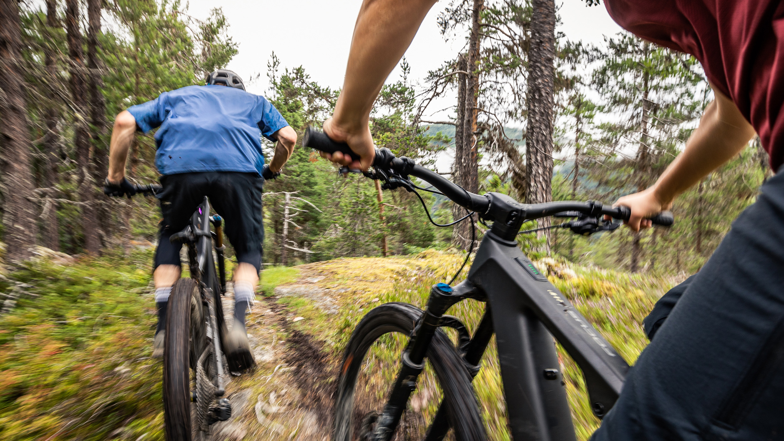 Looking at mountain biker descending a trail, through the arms and handlebar of a mountain biker following.