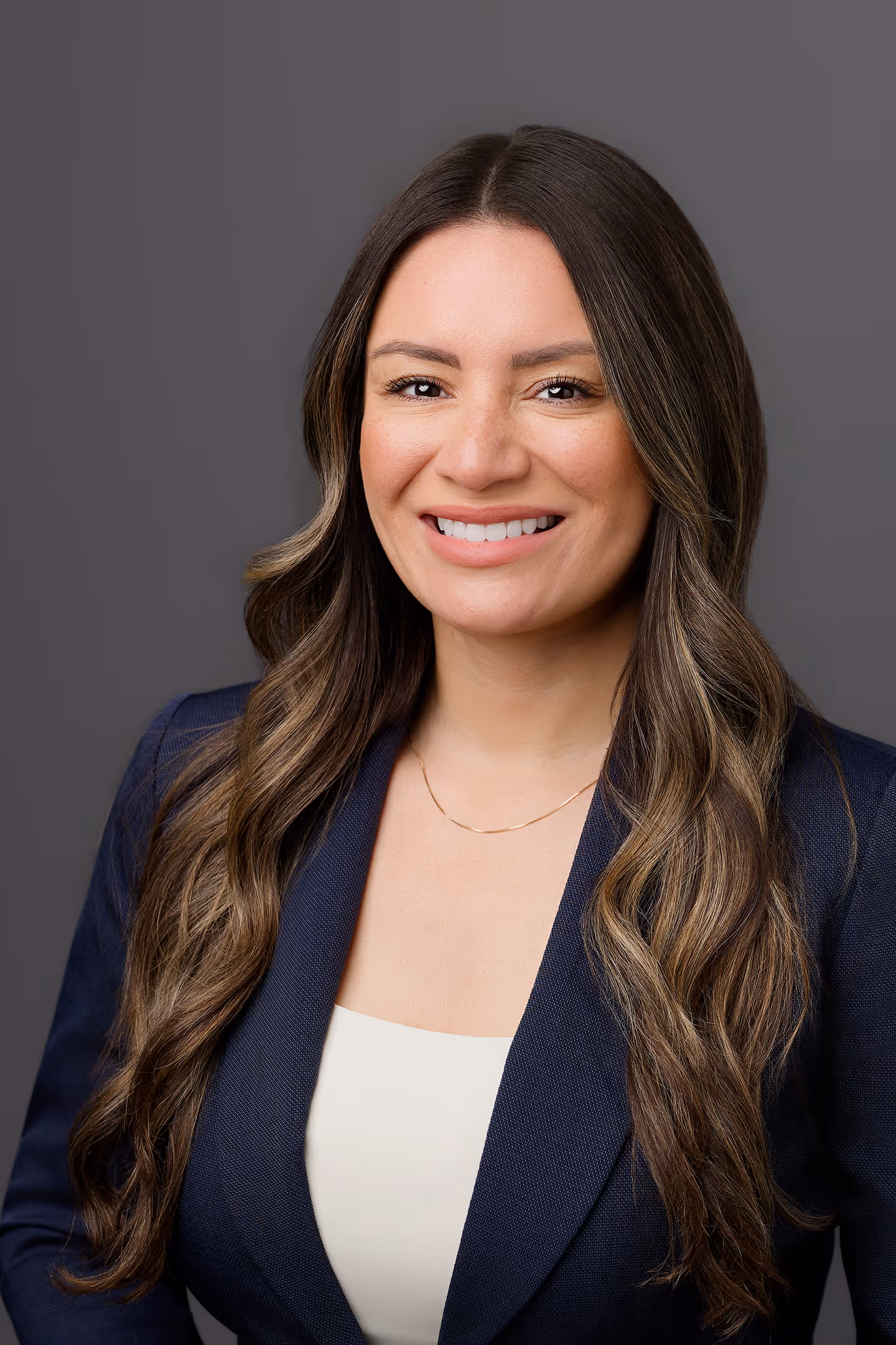Smiling woman with long wavy hair wearing navy blazer and white top against gray background.