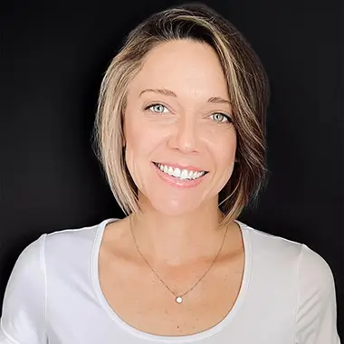 A woman with short, highlighted hair smiles warmly, wearing a white top and a delicate necklace, set against a dark background. She exudes friendliness.