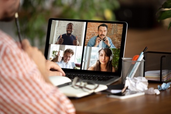 A person participates in a virtual meeting on a laptop, with four people visible on the screen. The desk has notebooks, glasses, and writing utensils.