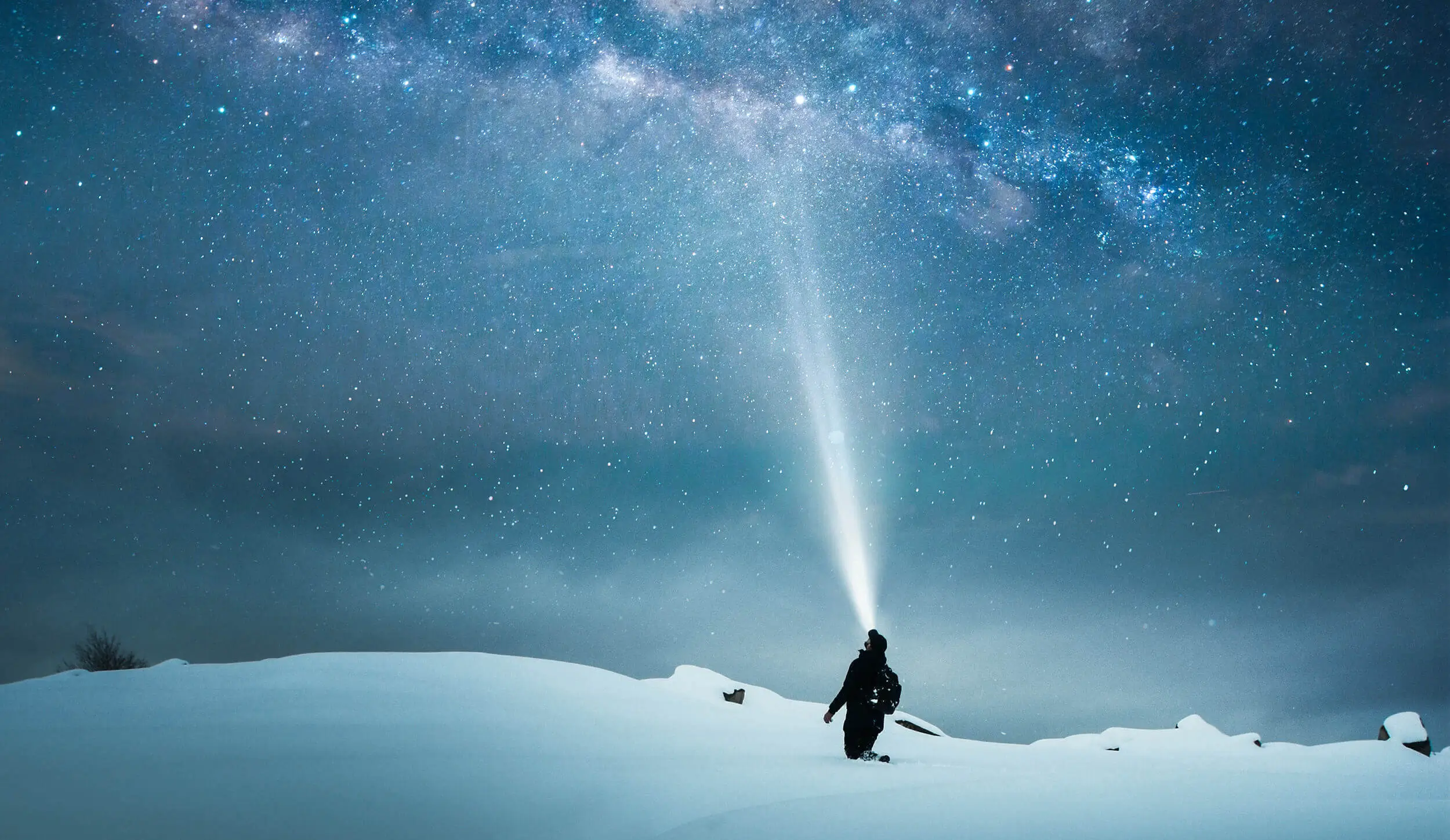 A person stands on a snowy landscape under a star-filled sky, shining a bright flashlight beam upward, merging with the cosmic display.
