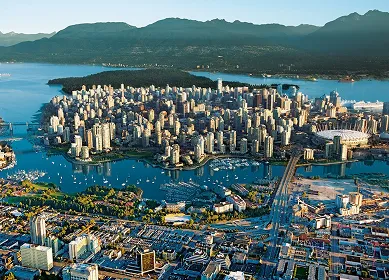 Aerial view of Vancouver cityscape featuring dense skyscrapers surrounded by water and mountains in the background.
