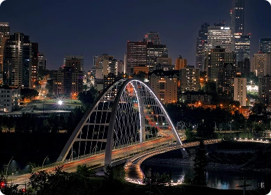 White arch bridge crossing a river with a city skyline in the background under a clear blue sky.
