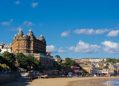 Sunny beach scene with a large historic hotel on the left and buildings along the coastline under a clear blue sky.