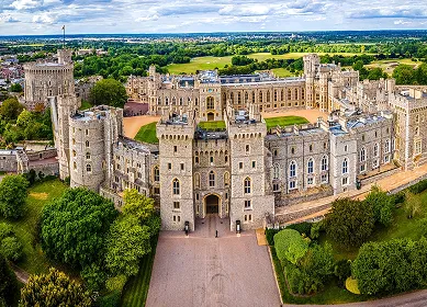 Aerial view of Windsor Castle, a large historic stone fortress with multiple towers and surrounded by green trees and gardens under a partly cloudy sky.