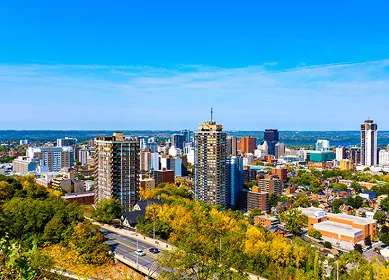 Cityscape of Hamilton with tall buildings, trees with autumn foliage, and a clear blue sky.