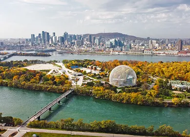 Aerial view of a park with a large geodesic dome near a river, autumn trees, and a city skyline in the background under a partly cloudy sky.