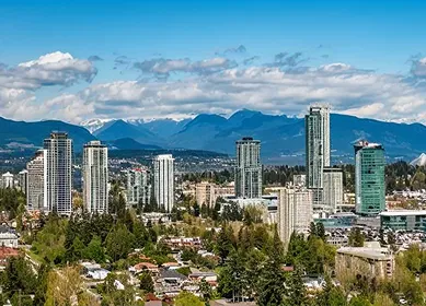 Cityscape with tall buildings surrounded by trees and mountains under a partly cloudy blue sky.