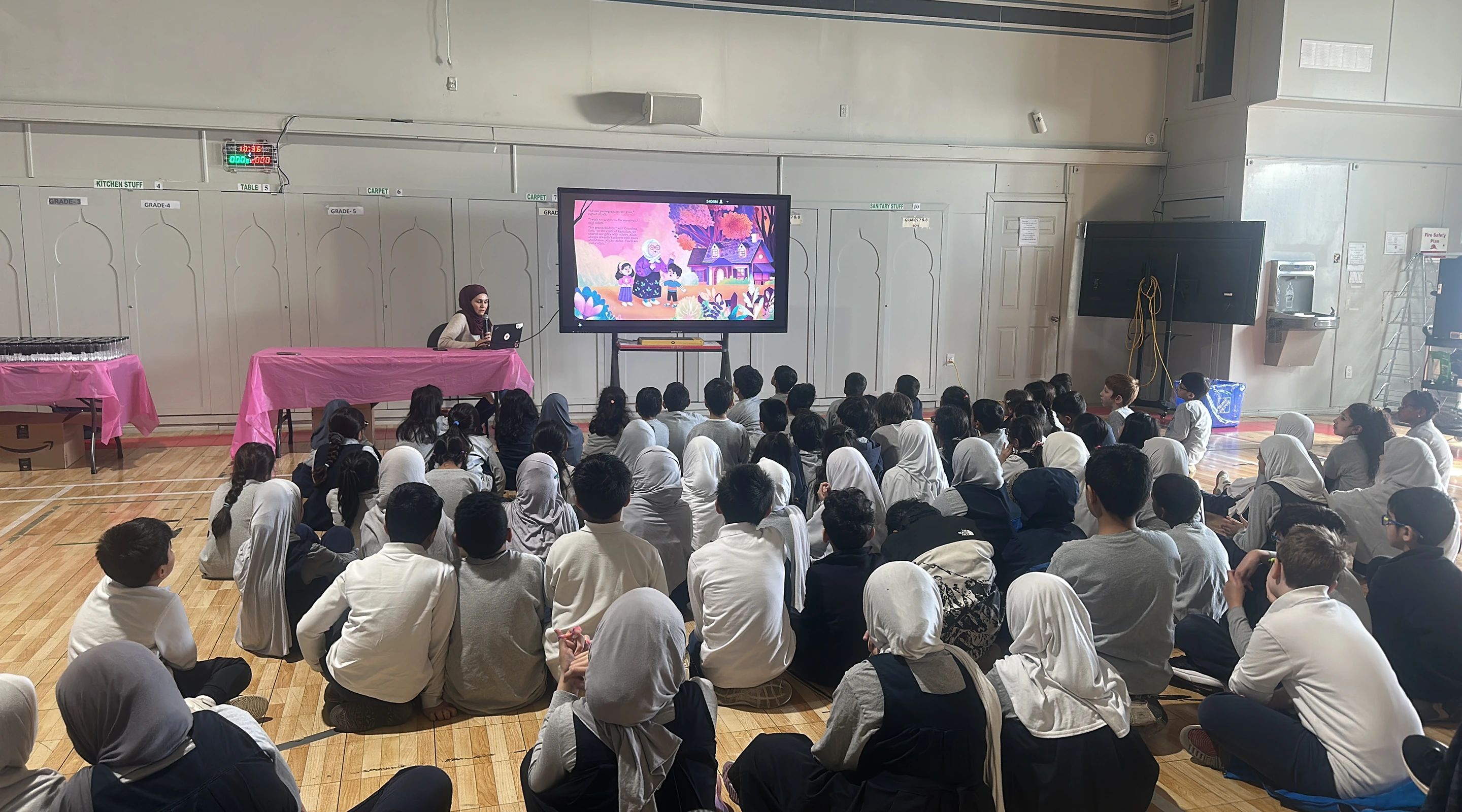A woman wearing a hijab addresses a large group of young students sitting on a gym floor while a storybook illustration is projected on a large screen.