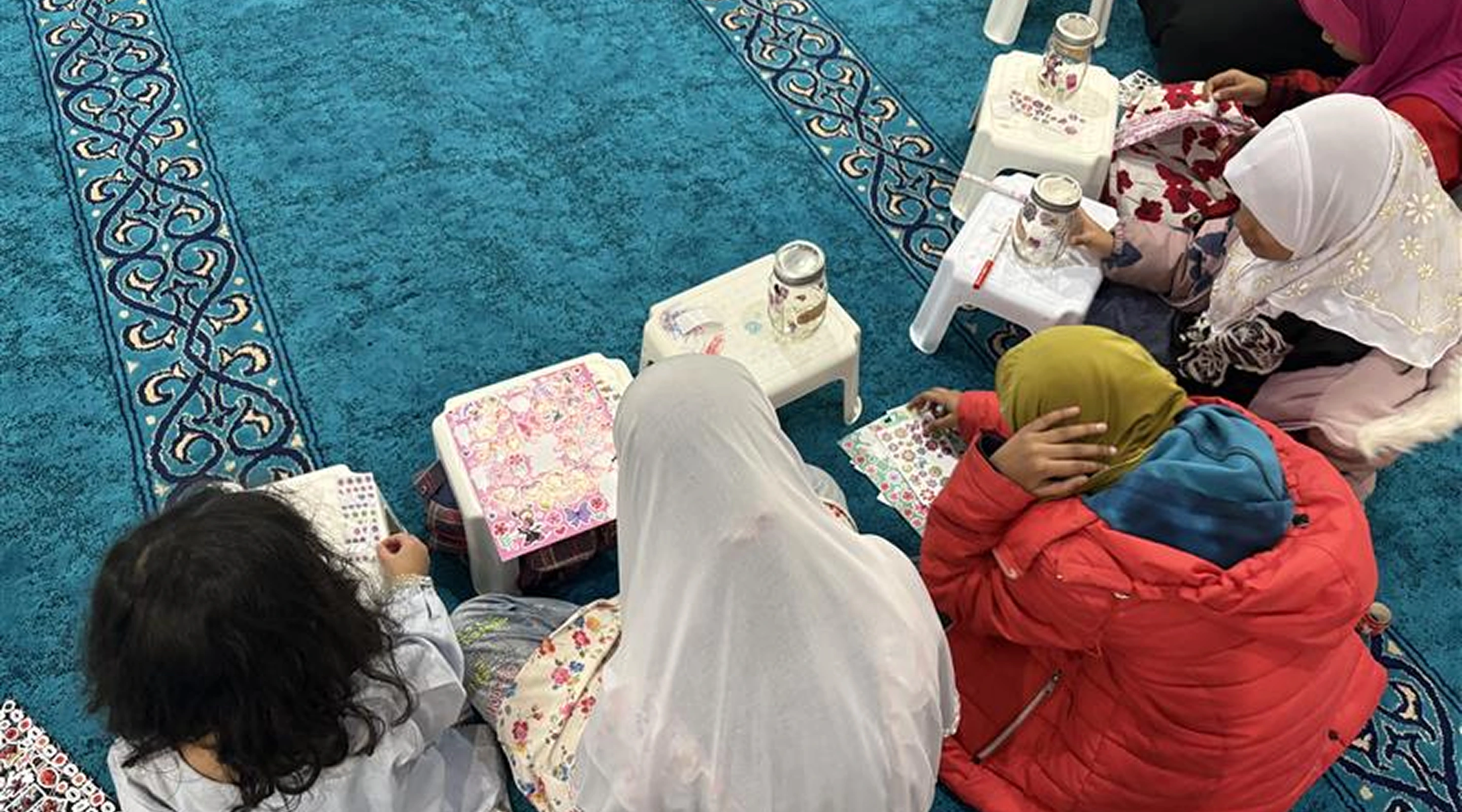 A high-angle view of a group of young girls sitting on a blue carpeted floor, working on arts and crafts with glass jars and colorful sticker sheets.