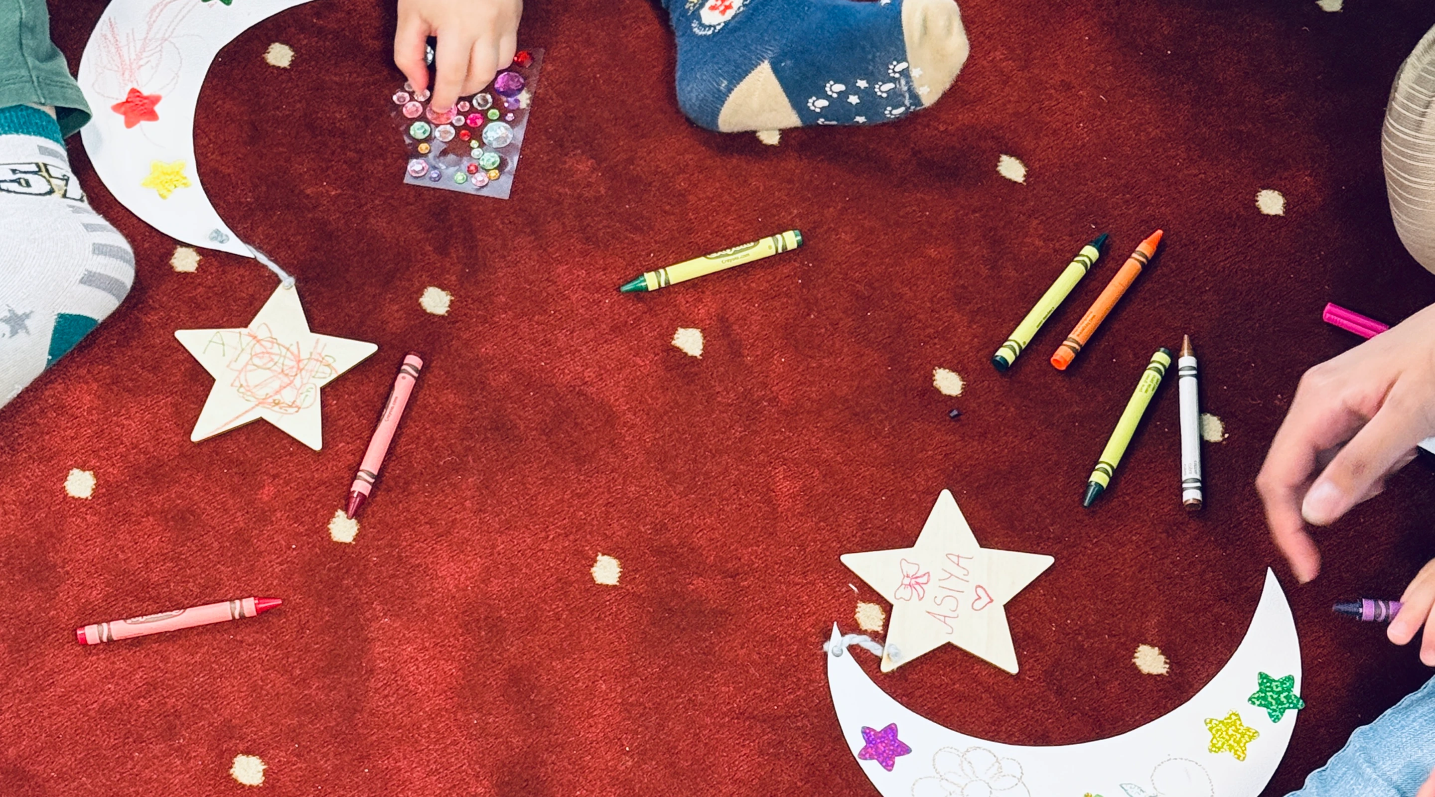 A top-down view of children sitting on a red patterned carpet making arts and crafts, featuring paper crescent moons and stars decorated with stickers and crayons.