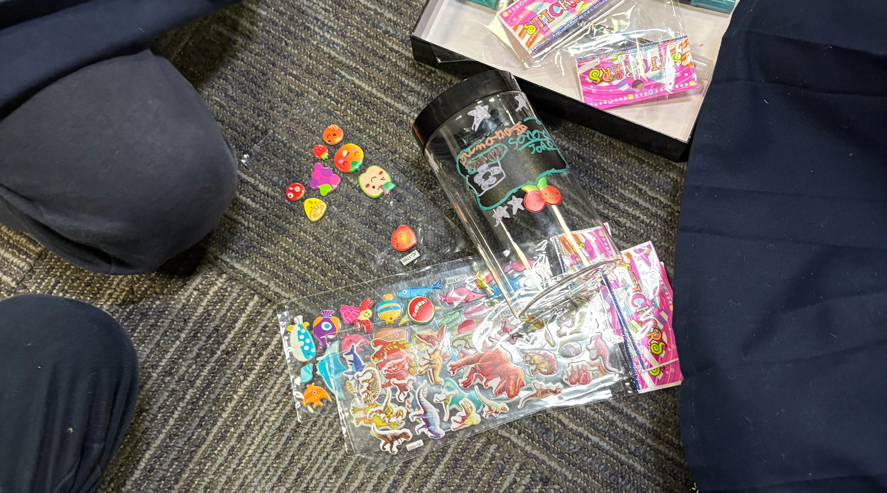 A top-down view of a clear plastic craft jar with a black lid resting on a grey carpeted floor alongside several packages of colorful stickers, including dinosaur and fruit designs.