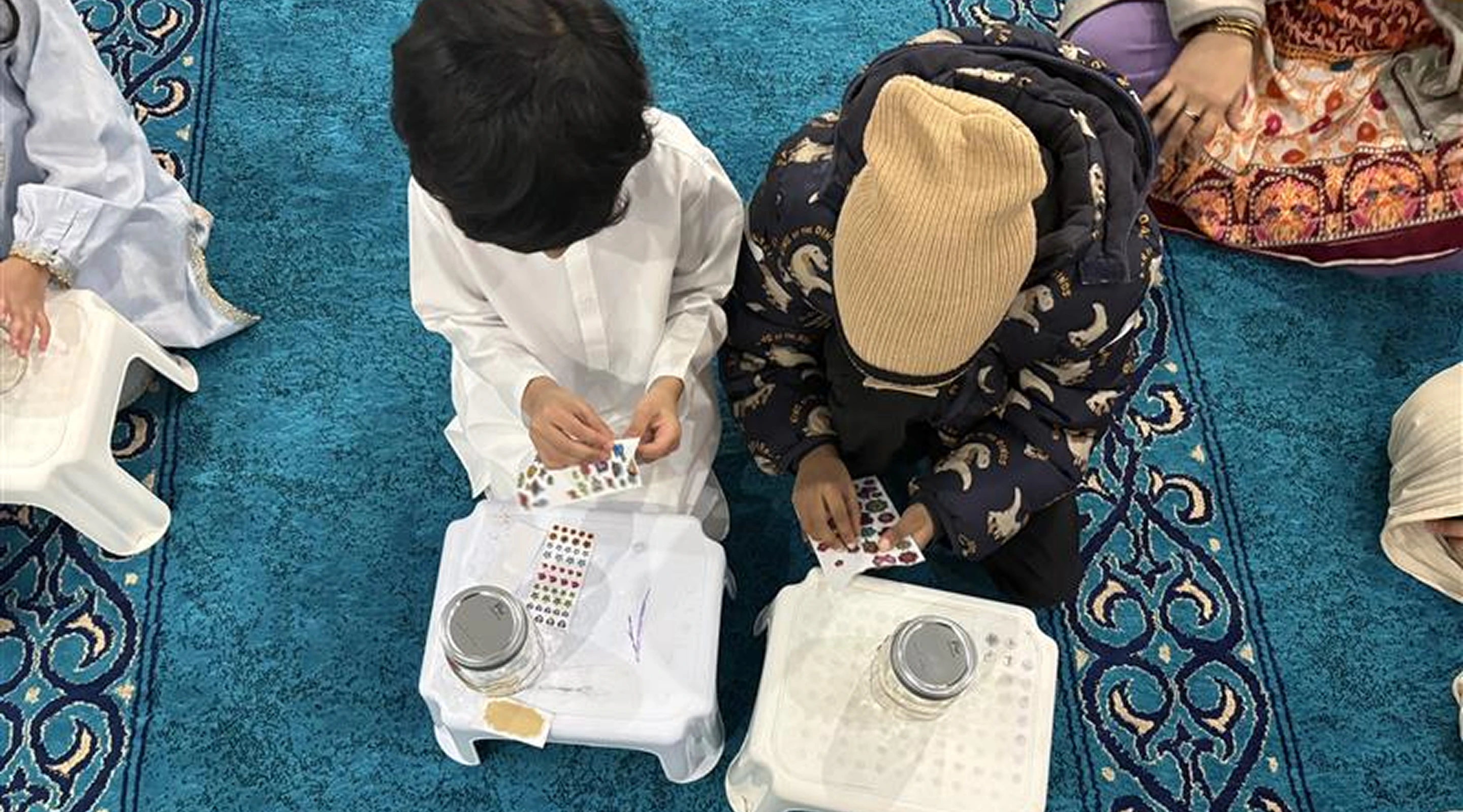A high-angle view of two young boys sitting on a blue patterned carpet, peeling stickers to decorate clear glass jars placed on small white stools.