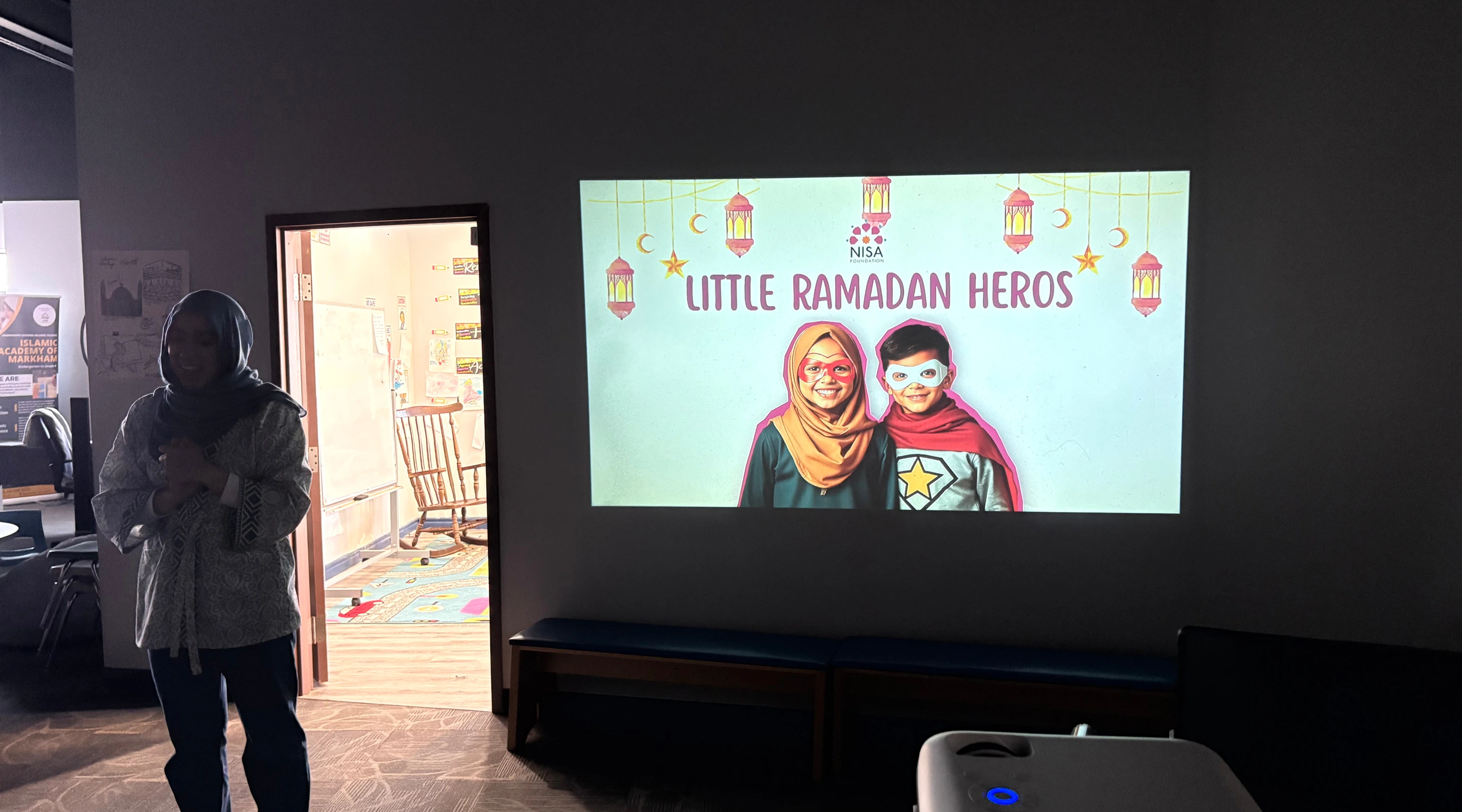 A woman wearing a hijab standing in a dimly lit room next to a bright projector screen displaying a presentation titled "LITTLE RAMADAN HEROS."