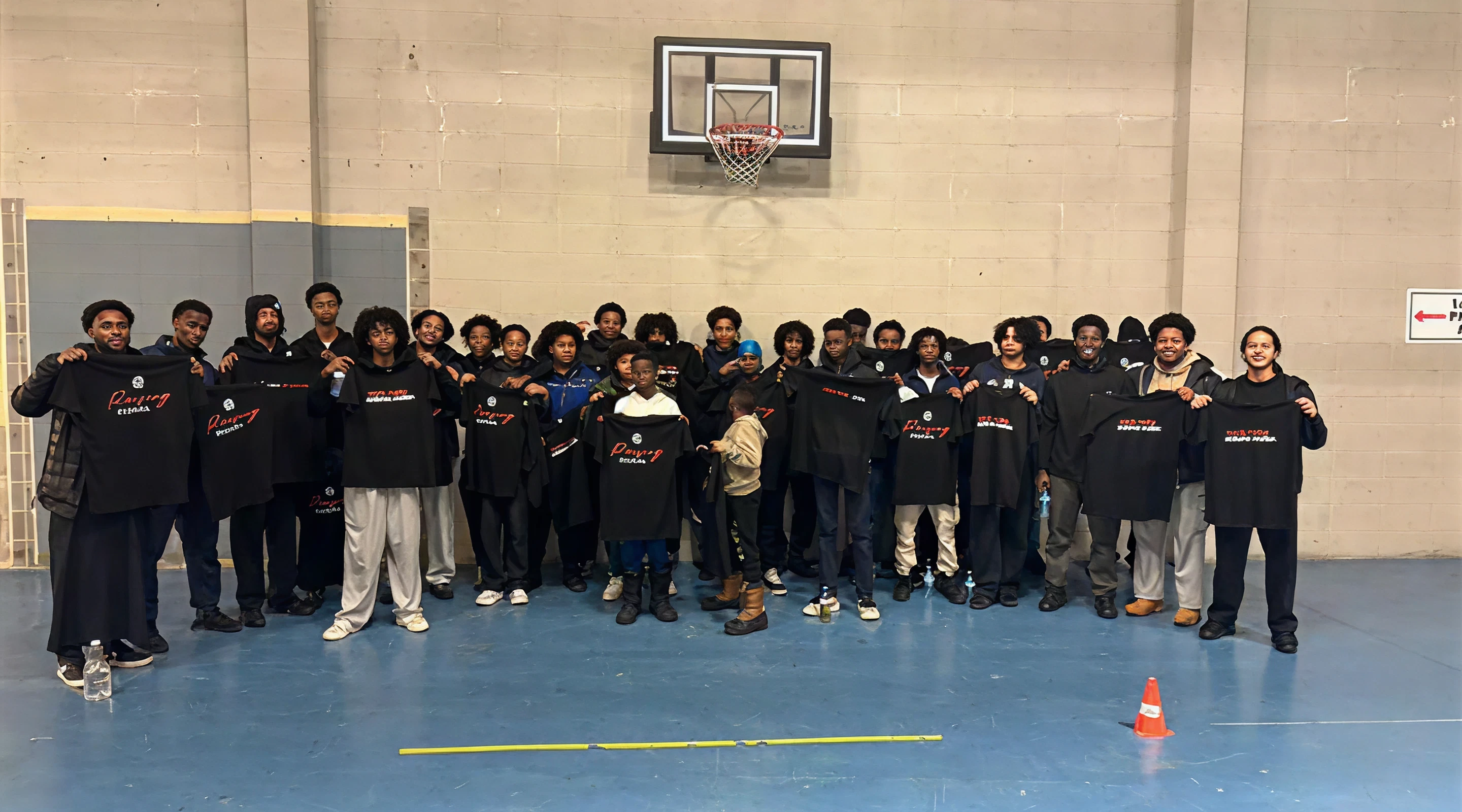 A large group of young Black men and boys standing together on an indoor basketball court, smiling and holding up matching black t-shirts with red and white text.