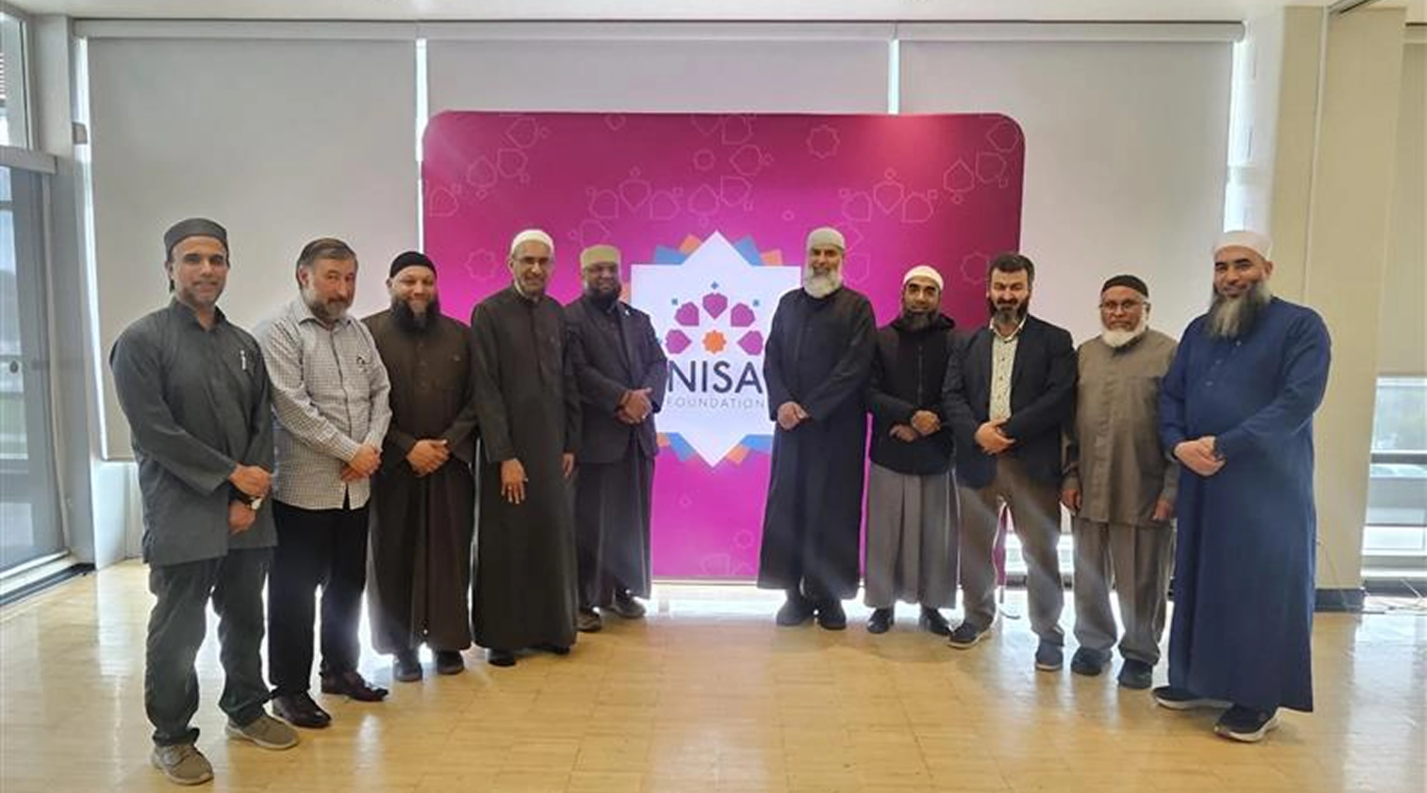 A group of ten men standing in a row and posing for a photo in front of a pink NISA Foundation backdrop.