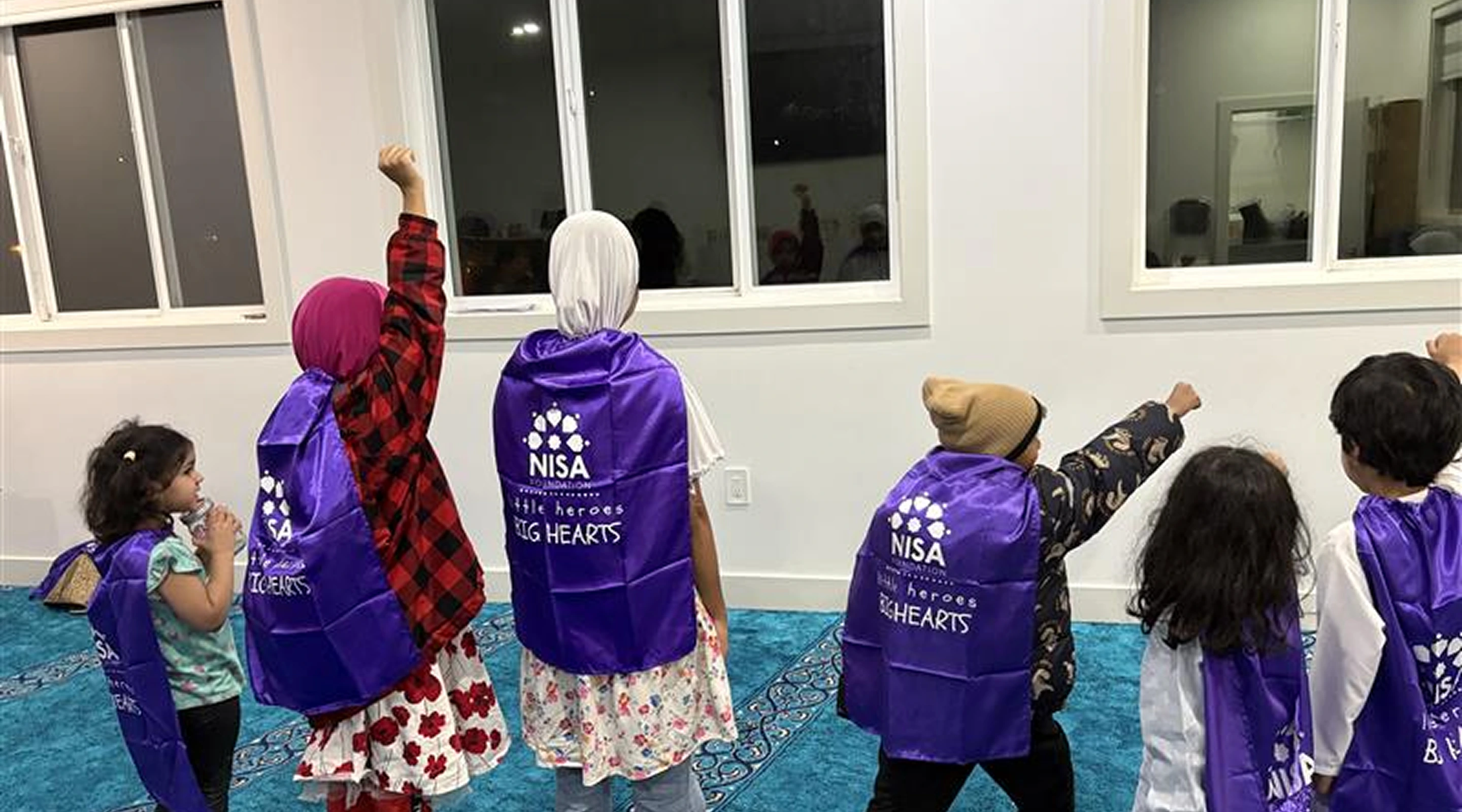 A group of young children wearing matching purple superhero capes standing on a blue carpeted floor and raising their fists in the air.