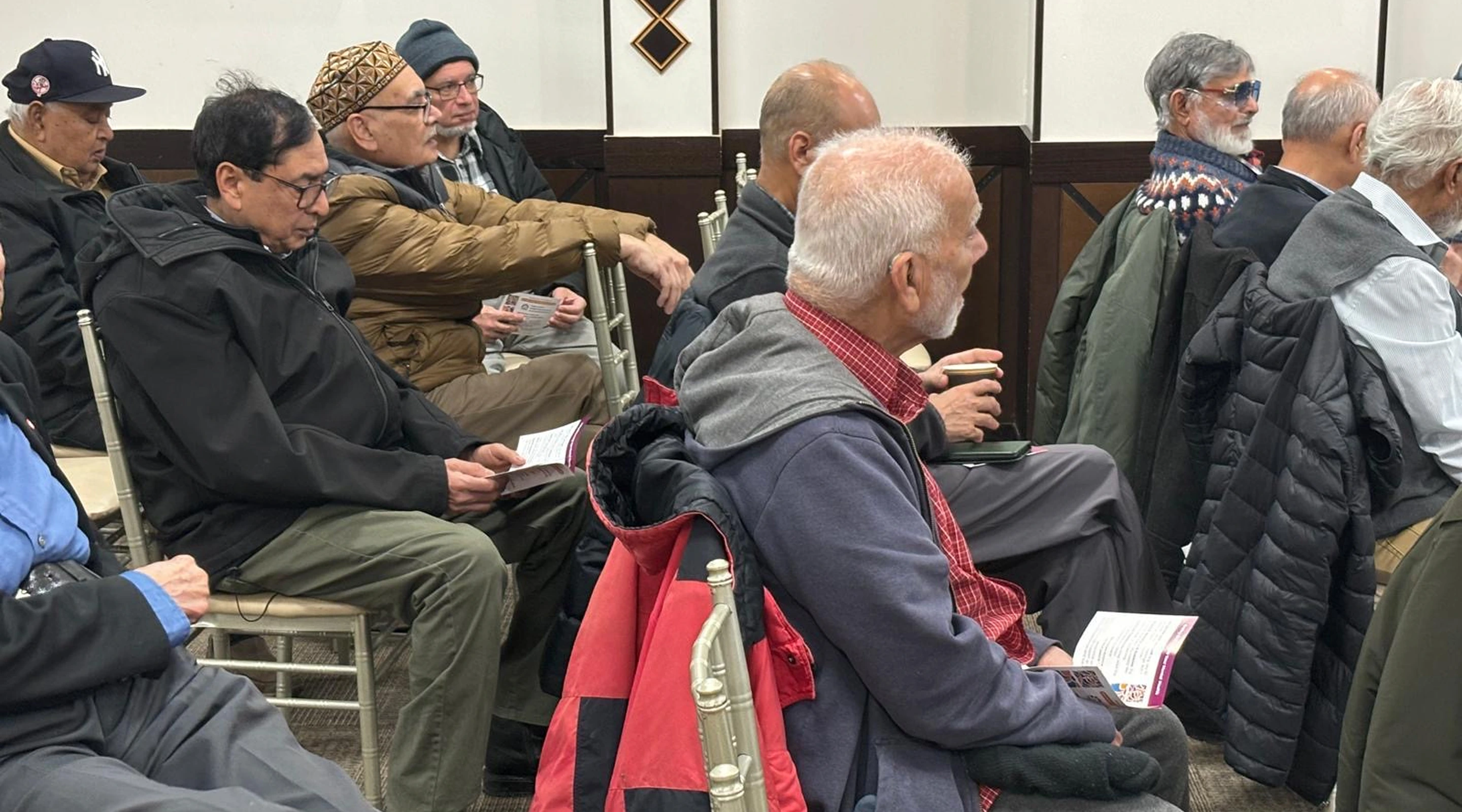 A side view of a group of older men seated in chairs, listening attentively and holding informational pamphlets during a community meeting.