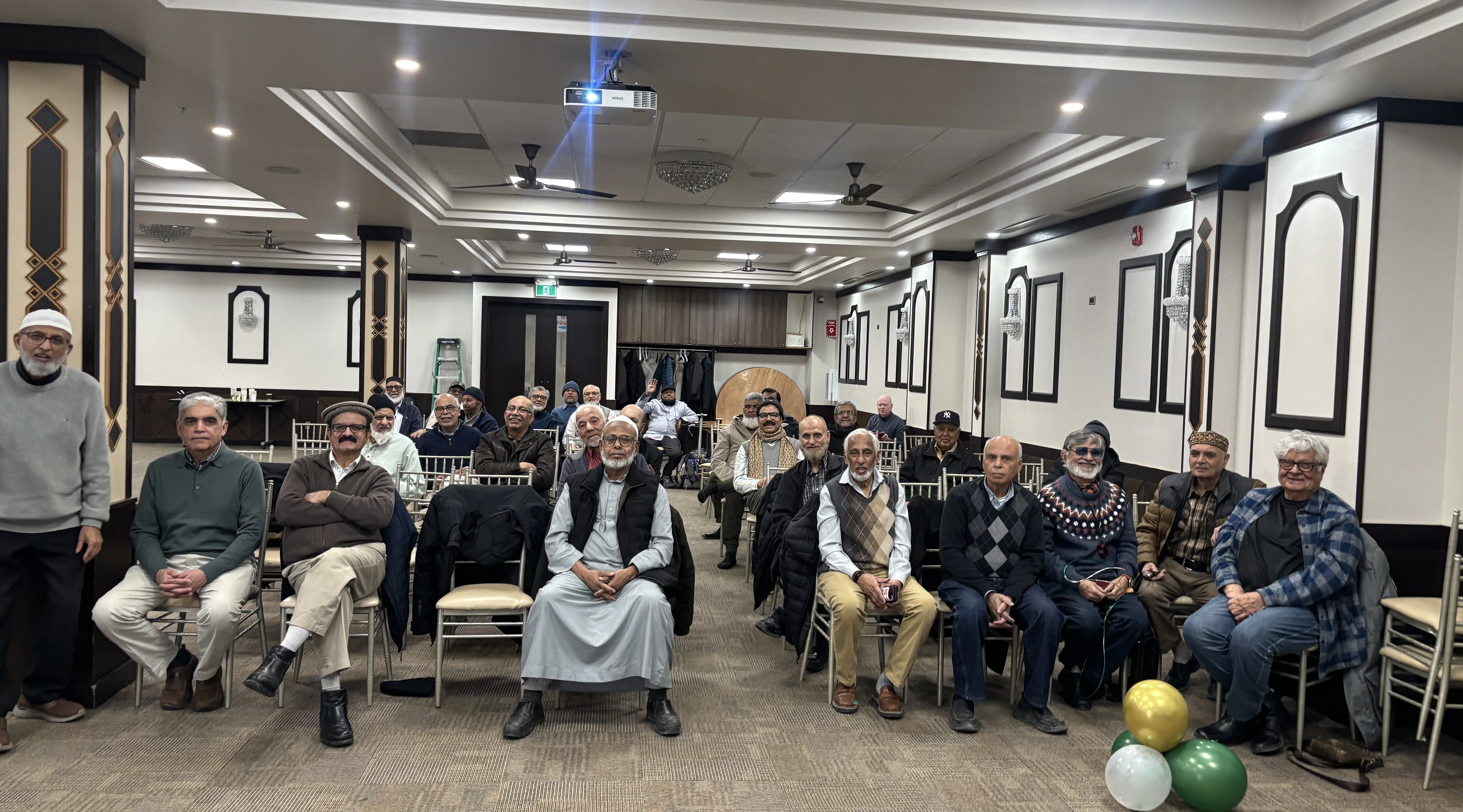 A group of older men sitting in rows of chairs during a gathering or community meeting inside a large, well-lit hall.