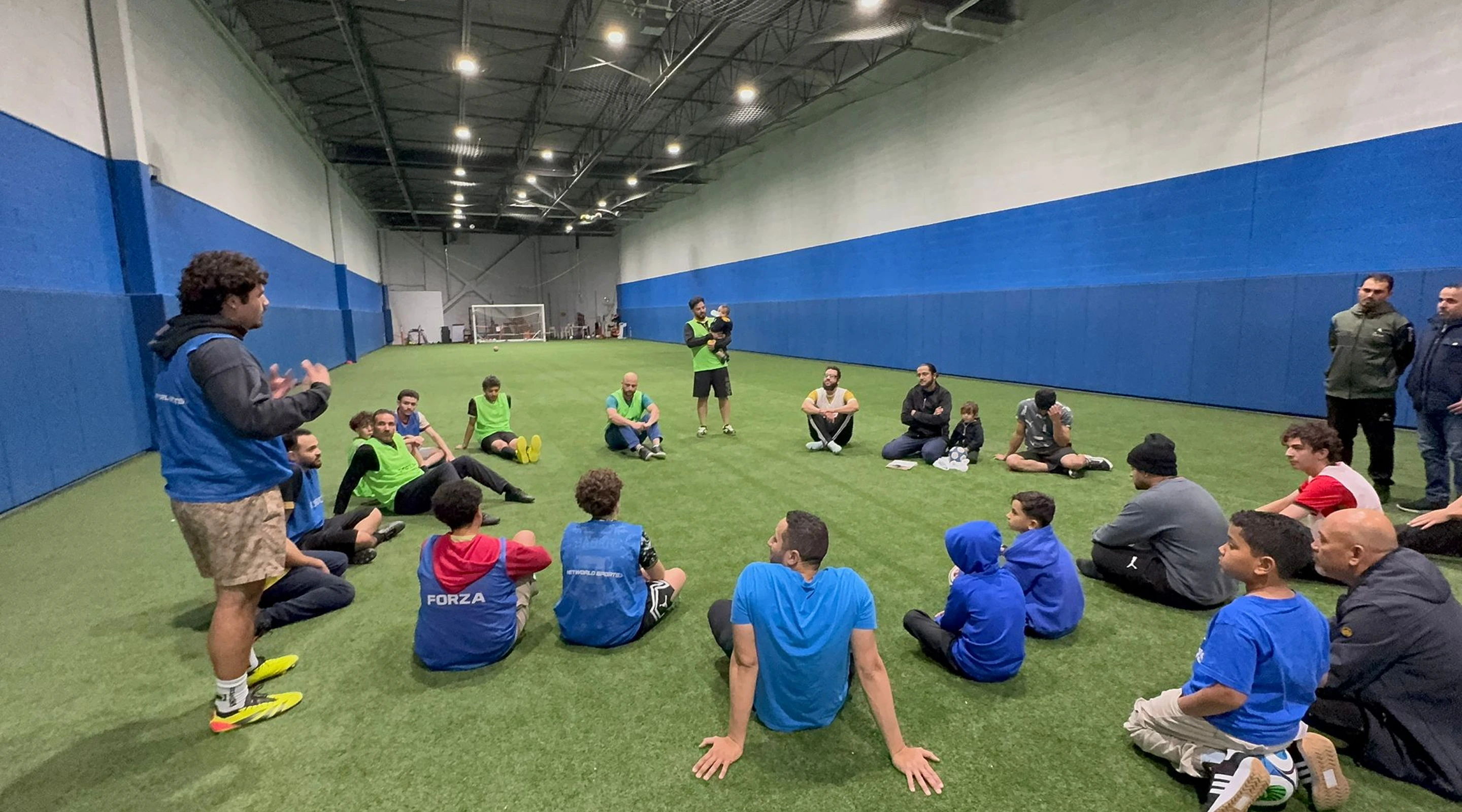 A coach or instructor standing and speaking to a group of men and children sitting in a large circle on an indoor green turf field.