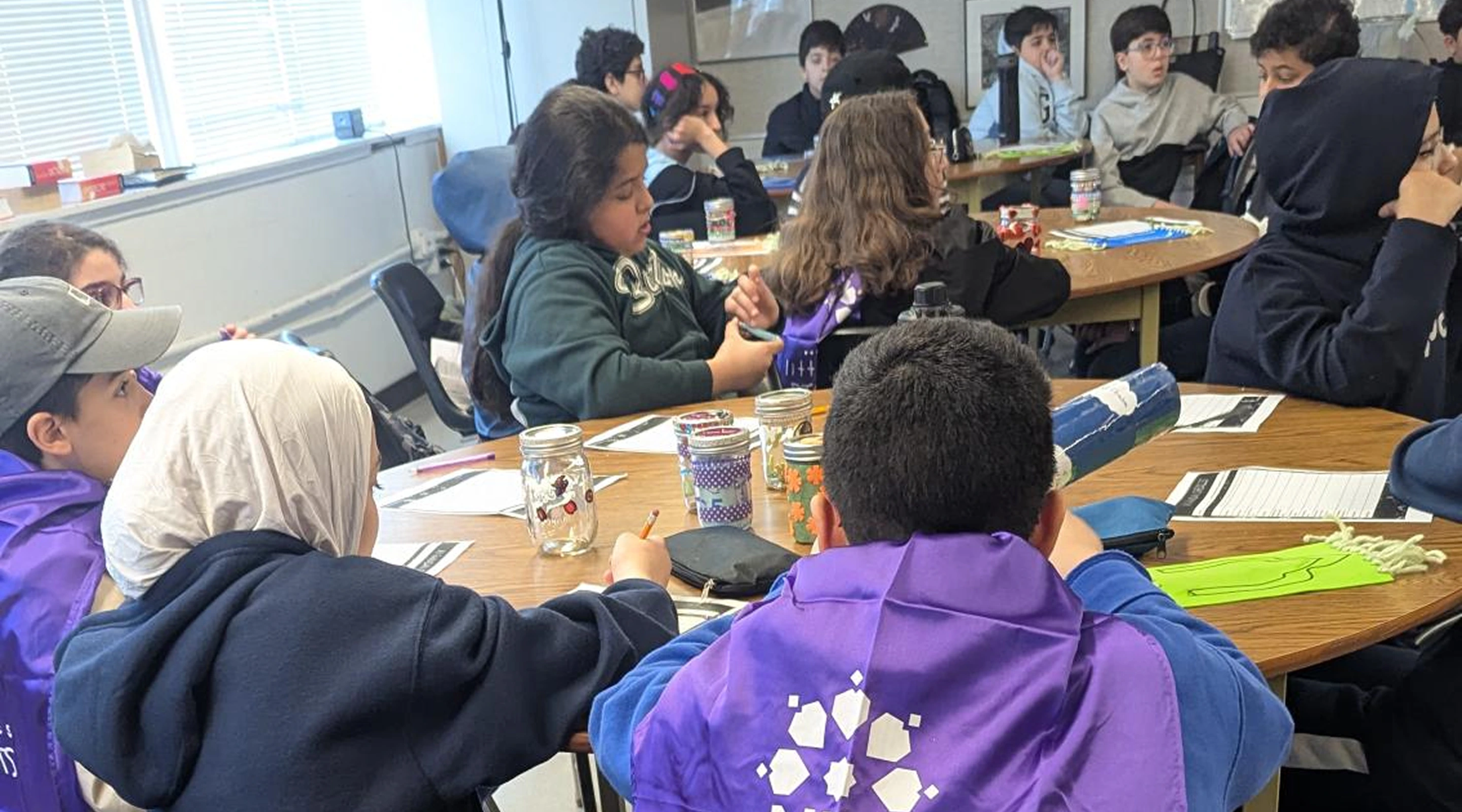 A group of young students sitting around large wooden tables in a bright classroom, working on crafts and activities with glass jars and papers.