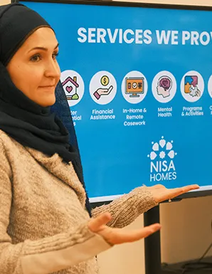 A woman in a dark hijab gesturing toward a blue presentation screen showcasing Nisa Homes services including financial assistance and mental health