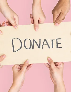 Diverse group of hands coming together from all sides to hold up a cardboard sign that says DONATE against a solid pink background