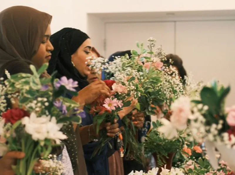 A group of women wearing hijabs participate in a floral arrangement workshop, carefully holding and assembling bouquets of colorful flowers and greenery.