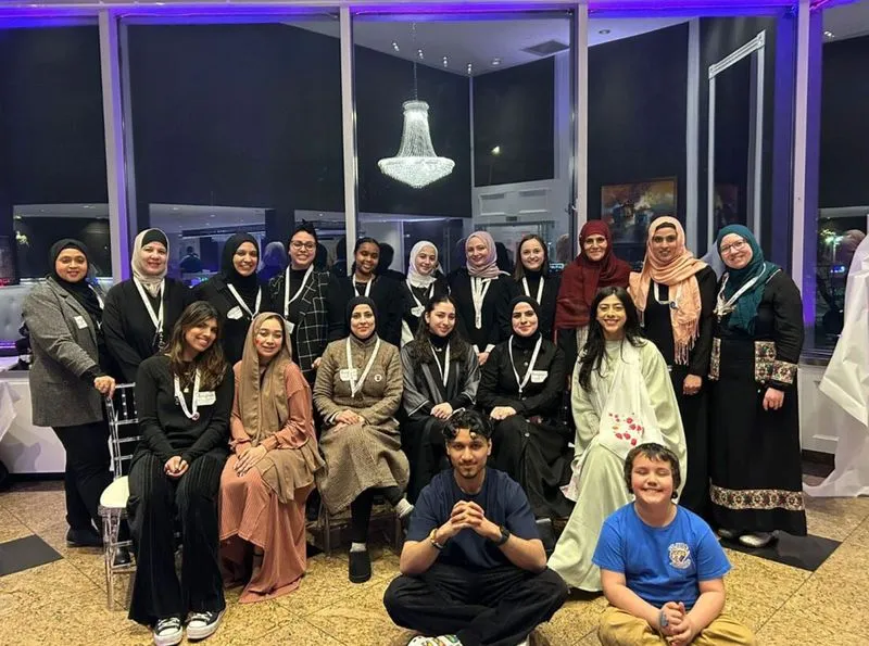 A large group of people, mostly women wearing hijabs, pose for a photo in a brightly lit room with floor-to-ceiling windows and a chandelier. A young man sits cross-legged in the front center, and a young boy sits next to him, while the rest of the group stands or sits in chairs behind them.