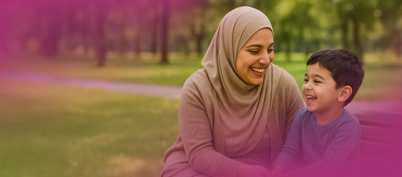 A woman wearing a beige hijab and sweater smiles warmly at a young boy who is laughing while sitting beside her in a park.