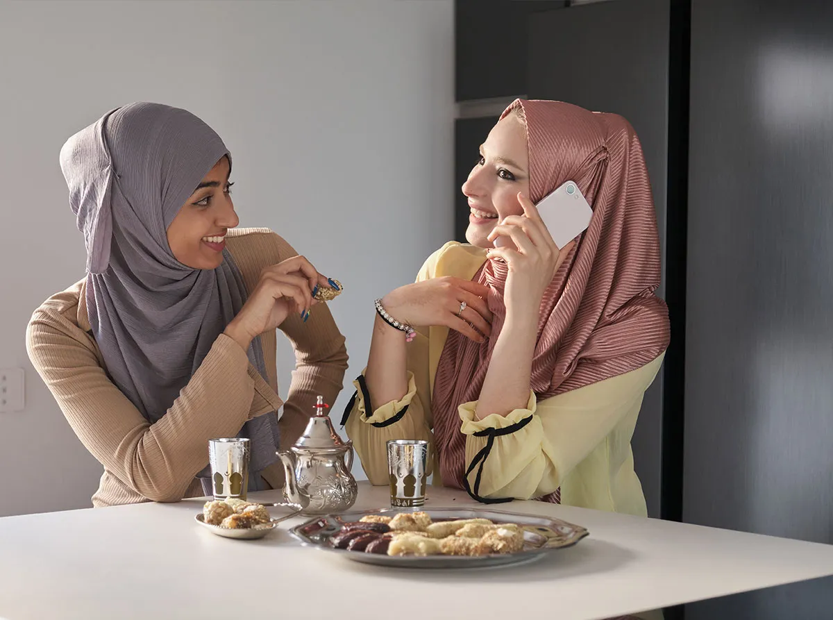Two smiling women wearing hijabs sit at a table with a silver teapot and plates of traditional pastries. One woman talks on a white smartphone while the other looks at her and holds a sweet.